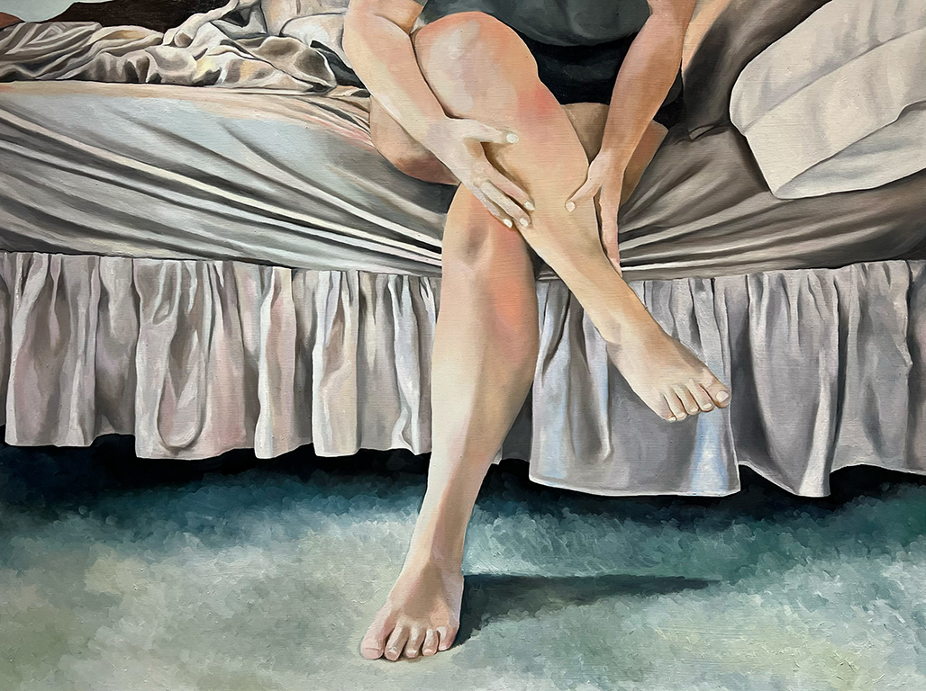 A person sitting on a bed, holding their leg with a relaxed posture, showcasing bare feet and a wrinkled bedspread.